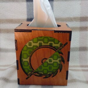 May include: A wooden tissue box cover with a green and brown geometric design. The design features a stylized saw blade.
