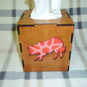 May include: A wooden tissue box cover with a pink and white geometric design cut out on the front. The box is made of stained wood and has a dark brown trim.