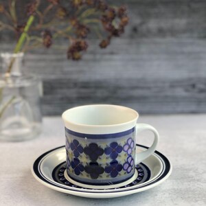 以下が含まれることがあります： 青と紫の花柄の白いセラミック製のコーヒーカップ。カップには持ち手があり、同じ柄の受け皿の上に置かれています。