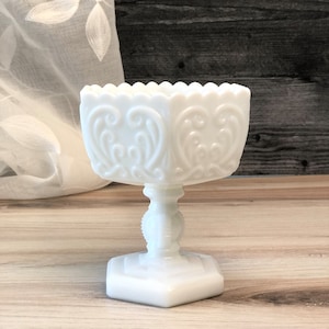 May include: White milk glass pedestal bowl with a scalloped edge and intricate floral design. The bowl is on a hexagonal base with a round pedestal.