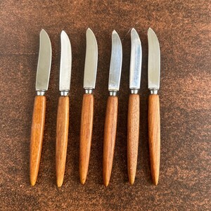 May include: Set of six butter knives with stainless steel blades and wooden handles. The knives are arranged in a row on a brown surface. The handles are a warm, medium brown color, and the blades are shiny silver.