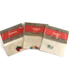May include: Three packages of iron-on appliques. The first package contains a red strawberry applique, the second package contains four heart appliques in different colors, and the third package contains a flower applique with three flowers in different colors. Each package has the text "Appliques Iron-On" and "Beachwood Ltd."