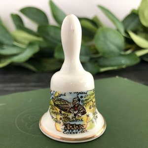 May include: A white ceramic bell with a gold trim. The bell is decorated with a map of England and features a small illustration of a person in a red and white costume. The text "England" appears twice on the bell.