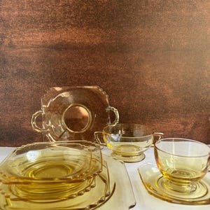 May include: A collection of vintage amber glass tableware, including a serving plate with handles, square plates, teacups, and saucers. The glassware has a scalloped edge design and is arranged on a white surface against a brown background.