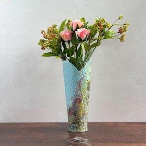 May include: A tall, light blue ceramic vase with a textured surface and small holes. The vase holds pink roses and green foliage. The vase has a unique, artistic design with colorful accents of red, yellow, and green.