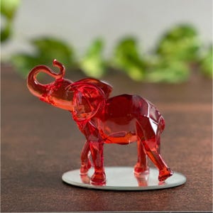 May include: A faceted, red glass elephant figurine stands on a small, round, mirrored base. The elephant has its trunk raised and is set against a blurred green and brown background. The figurine is a decorative item.