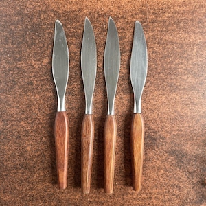 Könnte beinhalten: Vier Buttermesser mit Edelstahlklingen und Holzgriffen. Die Klingen haben eine abgerundete Form, und ein Messer hat eine gezahnte Kante. Die Griffe sind warmbraun, und die Messer sind in einer Reihe angeordnet.