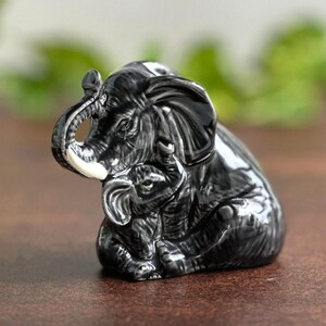 Könnte beinhalten: Glänzende, schwarze Keramikfigur eines erwachsenen Elefanten und eines Baby-Elefanten. Der erwachsene Elefant hat weiße Stoßzähne und umarmt den kleineren Elefanten. Die Figur steht auf einer braunen Oberfläche.