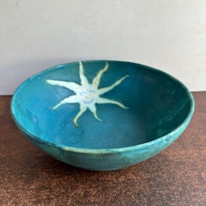 May include: A round ceramic bowl with a deep teal glaze and a hand-painted light cream sun motif at the center. The sun design features a simple face with eyes and a mouth, surrounded by eight wavy rays extending toward the bowl's curved rim.