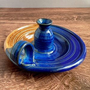 May include: A blue and brown ceramic dish with a small blue vase in the center. The dish has a unique handle that is shaped like a wave.
