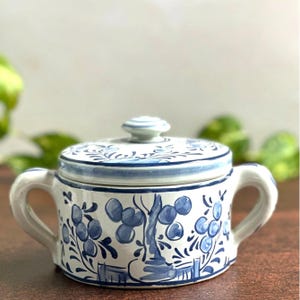 以下が含まれることがあります： 蓋と2つの取っ手が付いた白いセラミックの砂糖入れ。ボウルと蓋は、青い花柄と木の模様で装飾されています。蓋には小さな丸いノブが付いています。ボウルは茶色の表面にあります。