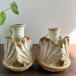 May include: Two ceramic candle holders with a unique design. Each holder features a cream-colored cup for the candle, surrounded by a brown and cream-colored petal-like structure. The holders sit on a round base with a brown rim.