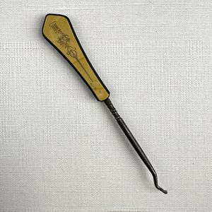 May include: An antique crochet hook with a decorative handle. The handle is a light tan color with black trim and etched designs. The hook itself is dark metal and has a curved end.