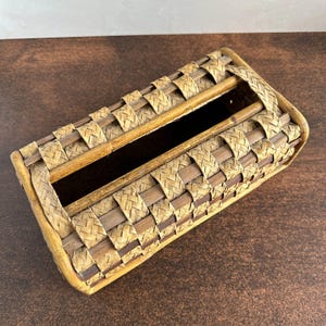 May include: A rectangular tissue box cover made of woven bamboo strips in shades of light and dark brown. The box has a rectangular opening on top and two wooden dowels for support. The woven pattern creates a textured surface.