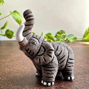 May include: A detailed, brown ceramic elephant figurine with a raised trunk and white tusks. The elephant has textured skin and decorative carvings. The figurine is a decorative item, suitable for home decor.