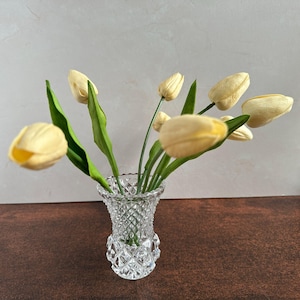 May include: A small, clear crystal vase filled with several pale yellow tulips and green leaves. The vase has a diamond-cut pattern. The tulips are in full bloom, creating a delicate floral arrangement.