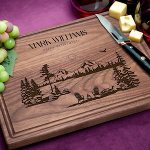 May include: Personalized walnut wood cutting board with a laser engraved design of a lake scene with mountains, trees, and two chairs. The text "Mark Williams Happy Retirement!" is engraved on the board.