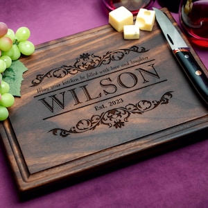 May include: A dark brown wooden cutting board with an engraved design and the text "May your kitchen be filled with love and laughter WILSON Est. 2023".
