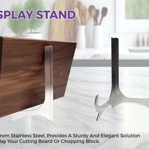May include: A silver-colored stainless steel display stand with a curved base and a vertical support arm. One stand is shown supporting a rectangular dark brown wooden cutting board, while a second identical stand is shown standing empty.
