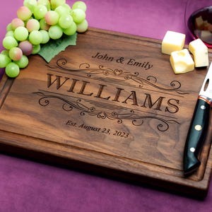 May include: Personalized walnut cutting board with a decorative engraved design. The board features the text "John & Emily Williams Est. August 23, 2023".