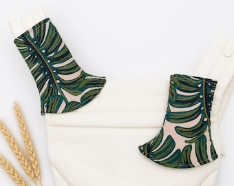 May include: A pair of cream-colored baby carrier strap covers featuring a dark green and light pink monstera leaf pattern. The covers are rectangular with rounded edges and are attached to the padded shoulder straps of a cream baby carrier.