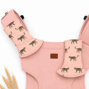 Puede incluir: Un portabebés rosa con detalles de estampado de leopardo. El portabebés tiene correas acolchadas para los hombros y un diseño de apoyo. El estampado de leopardo es un patrón repetido de manchas marrones sobre un fondo rosa claro. Una pequeña etiqueta de cuero marrón está cosida en la parte delantera.