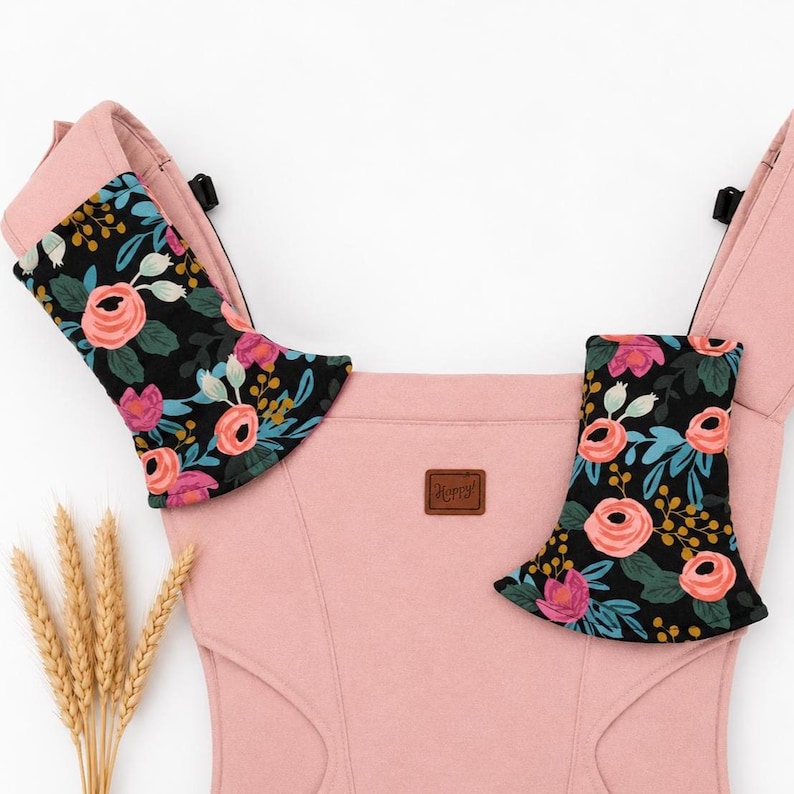 May include: A pink baby carrier with black floral print shoulder pads. The pads feature pink roses, blue leaves, and gold accents on a black background. A small brown leather tag with the word "Happy" is sewn on the front.