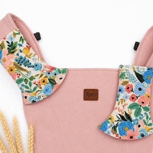 May include: A pink baby carrier with floral patterned accents. The carrier features a soft pink fabric with colorful floral designs in blue, pink, yellow, and green. A small brown leather tag reads "Happy!"
