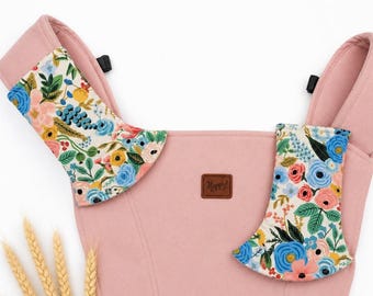May include: A pink baby carrier with floral patterned accents. The carrier features a soft pink fabric with colorful floral designs in blue, pink, yellow, and green. A small brown leather tag reads "Happy!"