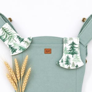 May include: A light green baby carrier with a forest print on the shoulder pads. The carrier has a brown leather tag with the brand name. Wheat stalks are in the foreground.