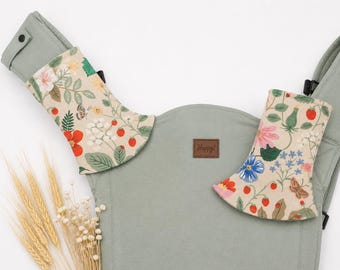 May include: A sage green baby carrier with floral patterned shoulder pads. The pads feature red, pink, and blue flowers, strawberries, and butterflies on a beige background. A brown leather tag reads "Happy!"