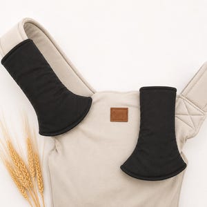 May include: A beige baby carrier with black shoulder strap covers. The carrier has a small brown leather patch and is set against a white background. A few stalks of wheat are in the lower left corner.