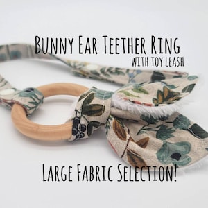 May include: A wooden teether ring with a fabric bow attached. The bow is made of a beige fabric with a floral pattern in shades of green, blue, and brown. The teether ring is attached to a fabric loop, which can be used as a toy leash. The text "Bunny Ear Teether Ring with Toy Leash" and "Large Fabric Selection!" is printed on the image.