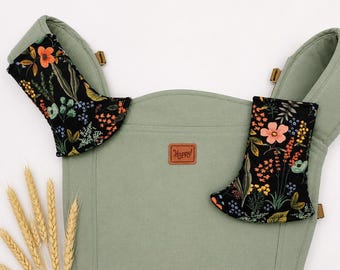 May include: A sage green baby carrier with black floral print accents. The carrier has a brown leather patch with the word "Happy!" and wheat stalks in the foreground. The floral print includes orange, pink, and yellow flowers.