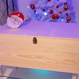 Pu&ograve; includere: Una scatola di legno per la conservazione con un coperchio incernierato e una chiusura in ottone. La scatola &egrave; appoggiata su un tavolo di vetro con un albero di Natale bianco sullo sfondo. Una statuetta di Babbo Natale &egrave; seduta sopra la scatola.