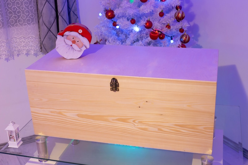 Pu&ograve; includere: Una scatola di legno per la conservazione con un coperchio incernierato e una chiusura in ottone. La scatola &egrave; appoggiata su un tavolo di vetro con un albero di Natale bianco sullo sfondo. Una statuetta di Babbo Natale &egrave; seduta sopra la scatola.