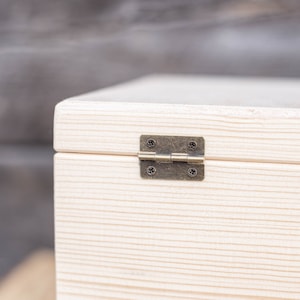 May include: Close-up of a white wooden box with a brass hinge. The box has a natural wood grain and is made of unfinished wood.