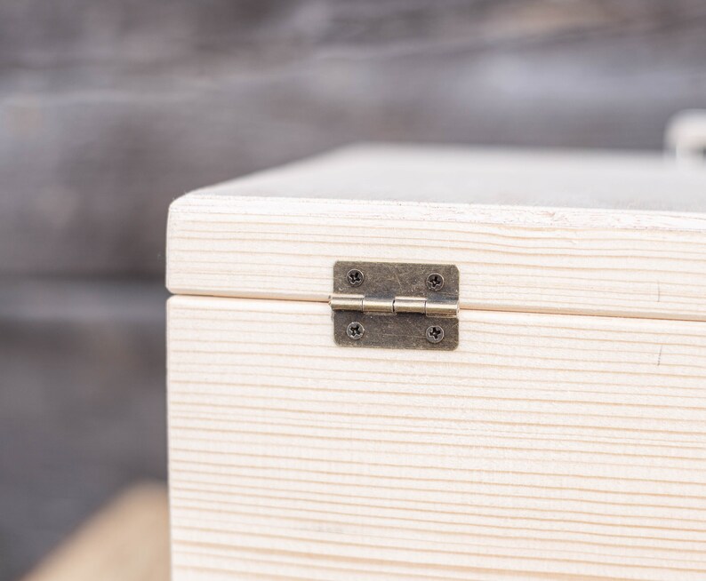 May include: Close-up of a white wooden box with a brass hinge. The box has a natural wood grain and is made of unfinished wood.