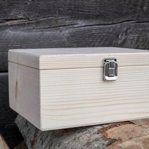 May include: A plain wooden box with a hinged lid and a silver latch. The box is sitting on a stack of tree rounds.