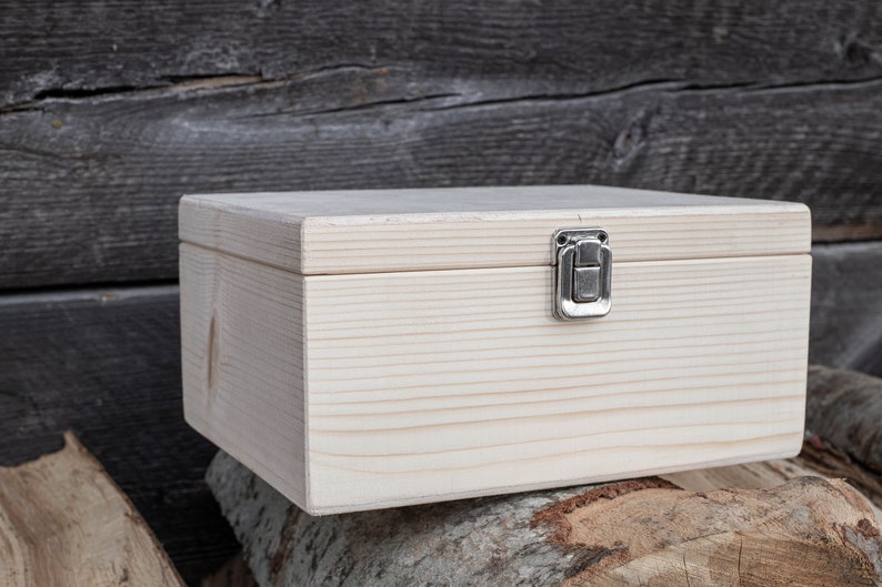May include: A plain wooden box with a hinged lid and a silver latch. The box is sitting on a stack of tree rounds.