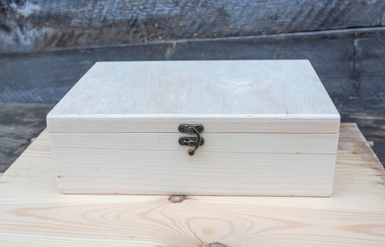 K&ouml;nnte beinhalten: Eine wei&szlig;e Holzkiste mit einem Messingverschluss. Die Kiste steht auf einer hellen Holzoberfl&auml;che.