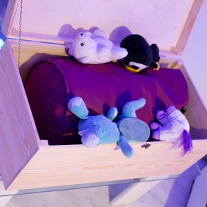 Pu&ograve; includere: Un baule di legno per giocattoli con una fodera viola e una variet&agrave; di giocattoli di peluche all'interno, tra cui un pinguino, un unicorno e un dinosauro blu.