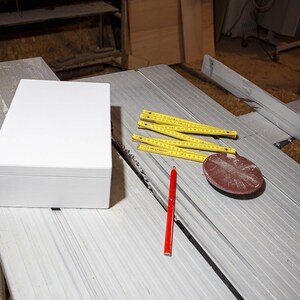 K&ouml;nnte beinhalten: Eine wei&szlig;e Box, ein roter Bleistift, ein gelbes Ma&szlig;band und eine braune Schleifscheibe auf einer grauen Werkbank.