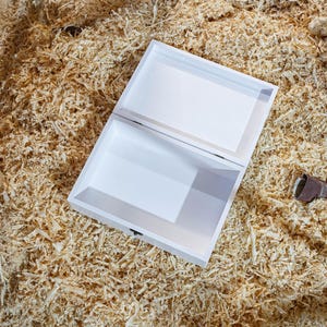 K&ouml;nnte beinhalten: Eine wei&szlig;e Holzkiste mit einem Scharnierdeckel ist ge&ouml;ffnet und zeigt ein leeres Inneres. Die Kiste steht auf einem Bett aus Holzsp&auml;nen.