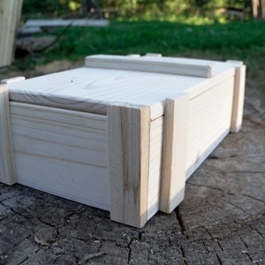 May include: A plain wooden box with a hinged lid. The box is made of light-colored wood and has a simple, rectangular shape.
