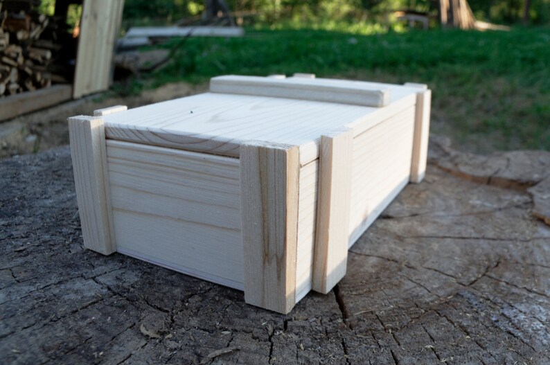 May include: A plain wooden box with a hinged lid. The box is made of light-colored wood and has a simple, rectangular shape.
