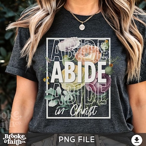 Può includere: Una maglietta grigia scura con un disegno floreale e il testo "ABIDE in Christ" in bianco.