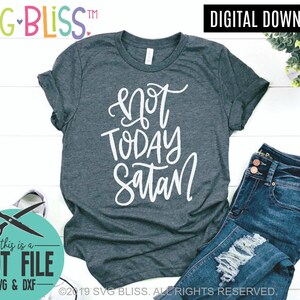 May include: A dark gray t-shirt with the text "not today satan" written in white cursive lettering. The shirt is laid flat on a white wooden surface with a pair of blue jeans and a pair of scissors in a circle with the text "this is a cut file -SVG & DXF- ".