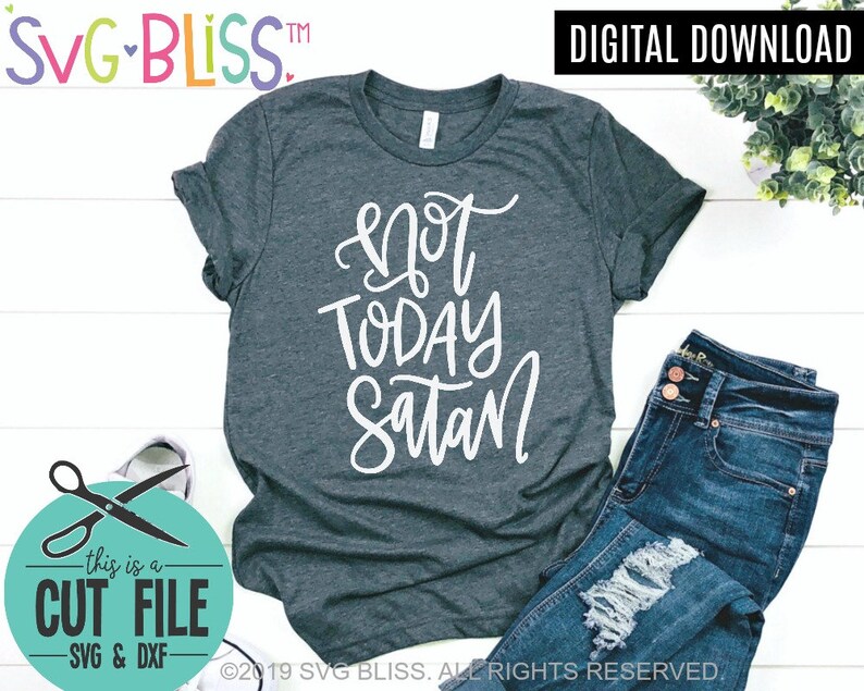 May include: A dark gray t-shirt with the text "not today satan" written in white cursive lettering. The shirt is laid flat on a white wooden surface with a pair of blue jeans and a pair of scissors in a circle with the text "this is a cut file -SVG & DXF- ".