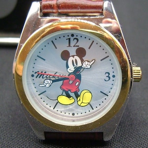May include: A silver and gold wristwatch with a brown leather band. The watch face features a cartoon image of Mickey Mouse with the word "Mickey" printed below. The watch face has black hour and minute hands and black Roman numeral hour markers.